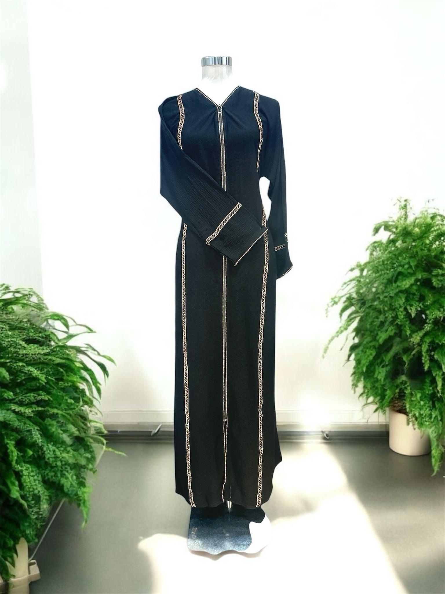 Our Nura Abaya features a sleek front opening with a central zip closure, framed by fine gold accents that elongate the figure and create a polished finish. Vertical trim panels along the body add structure and definition, while the subtly textured fabric enhances its overall elegance.