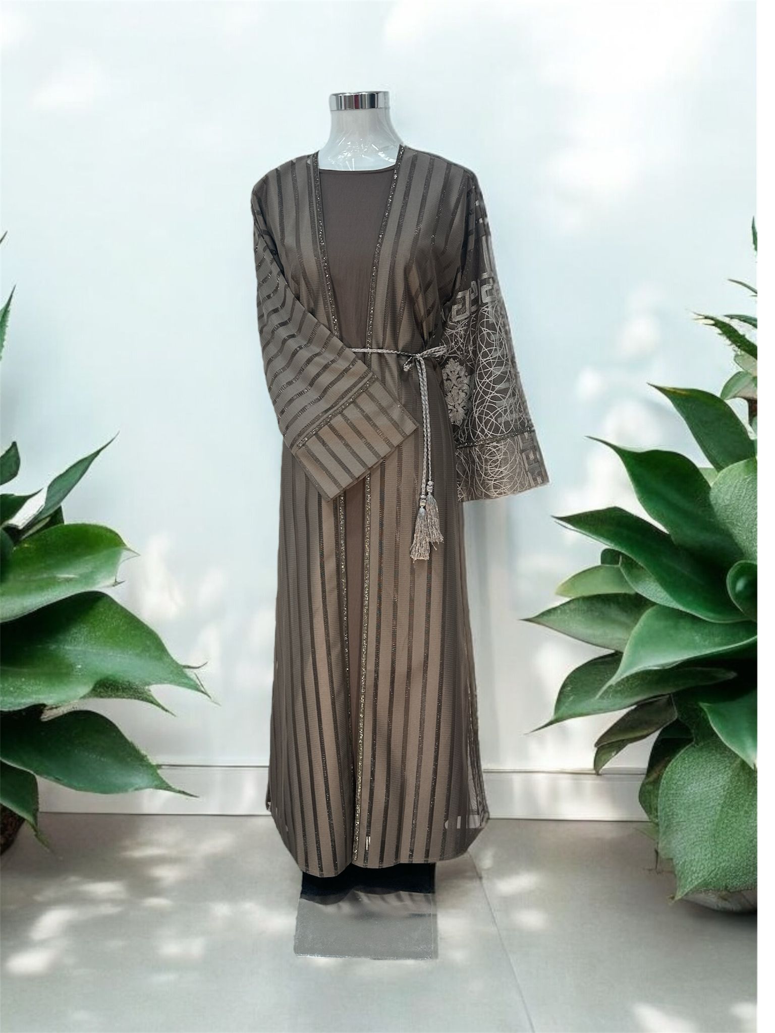 Step into effortless elegance with this sophisticated mocha-toned abaya, featuring shimmering vertical stripe detailing for a sleek and elongated silhouette. Designed with unique contrast sleeves showcasing intricate geometric and floral-inspired patterns, this piece blends modern style with timeless modest fashion. Complete with a matching inner dress and braided tassel belt, it’s perfect for both formal occasions and everyday elegance.